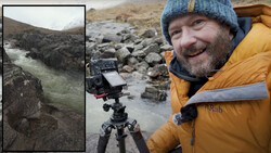 Filmmaker smiling at camera while operating cinema camera on tripod beside rushing glacial river in rocky mountain valley.