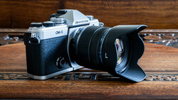 OM System OM-3 film camera with black leather grip and attached wide-angle lens with hood on wooden surface.