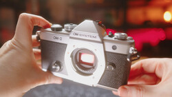 OM System OM-3 film camera held in hands against warm-toned blurred background.