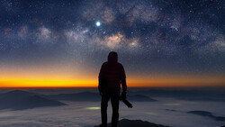 Silhouetted photographer standing on a mountain peak overlooking clouds at sunrise, with the Milky Way and stars visible in the night sky above.