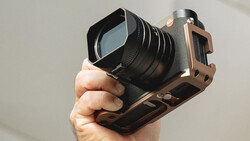 Hand holding a mirrorless camera with a square cage rig and telephoto lens against a neutral background.