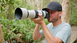 Photographer holding a telephoto lens camera at eye level outdoors among greenery.