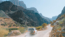 White SUV driving on a dusty mountain road through a dramatic canyon with steep rocky cliffs.