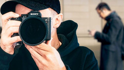 Photographer holding Sony mirrorless camera up to face while wearing black cap and jacket.