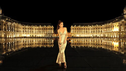 Woman in an elegant beaded gown standing centered in a symmetrical plaza reflected in still water at night.