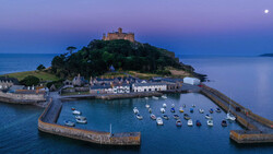Coastal village with castle on hilltop at twilight, moored boats in harbor below.