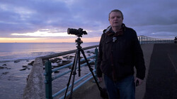 Photographer with camera on tripod at coastal location during twilight.