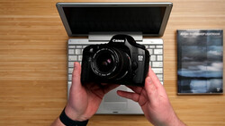 Canon EOS 5D Mark IV DSLR camera held above a laptop keyboard on a wooden desk.