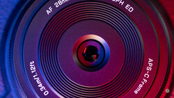 Close-up of a camera lens front element with concentric focus rings and text markings, illuminated by blue and red lighting.