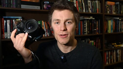 Man holding a mirrorless camera with a prime lens, speaking toward the camera in a home studio setting.