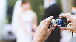 Hands holding a compact digital camera capturing a blurred outdoor event with people in the background.