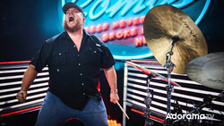 Drummer performing on stage with energetic expression and raised cymbals, vibrant neon backdrop.