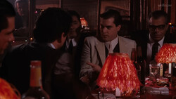 Men in suits gathered around a dinner table with red lampshades and glassware, engaged in conversation during an evening event.