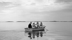 Four people sitting in a small rowboat on calm, glassy water under an overcast sky.