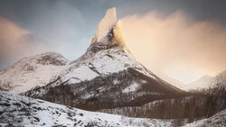 Snow-covered mountain peak with forested slopes under dramatic cloudy sky.