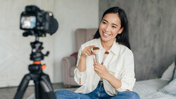 Content creator holding makeup brushes while seated in front of a camera on tripod setup.