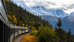 Passenger train curving through forested mountain landscape with snow-capped peaks in background.