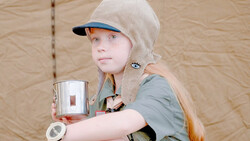 Young child wearing a cream and blue cap and olive shirt holds a paint can against a tan canvas backdrop.