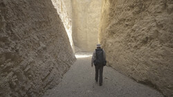 Lone figure walking away through a narrow alleyway between high earthen or mud-brick walls.