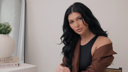 Woman with long black hair wearing a brown and black layered jacket, photographed indoors against a neutral wall.