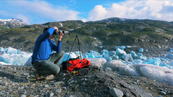 Photographer crouching with camera on tripod, documenting blue glacier icebergs with mountains in background.