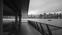 Black and white view from a covered waterfront pier toward a distant city skyline across calm water.