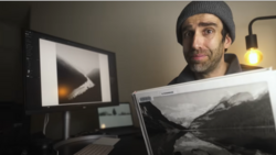 Photographer holding a landscape print while reviewing images on dual monitors in a studio workspace.