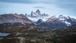 Snow-capped mountain peaks with jagged granite formations rising above a turquoise glacial lake and forested foothills.