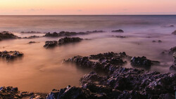 Rocky coastline at sunset with smooth water and layered rock formations shrouded in mist.
