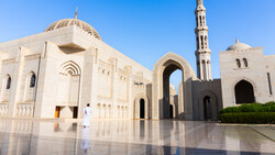 Sunlit courtyard of a mosque with cream-colored stone architecture, featuring a domed structure, tall minaret, and reflective polished floor.