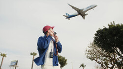 Man in blue jacket and maroon cap watches airplane flying overhead while holding binoculars to his face.