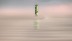 Green and yellow lighthouse emerging from thick fog over calm water.