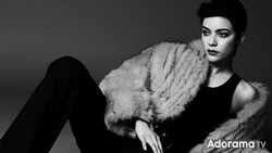 Person in black sleeveless top and fur stole against neutral background, gazing downward.