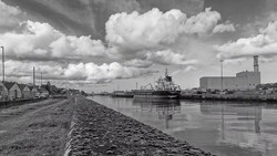 Industrial cargo ship moored alongside a stone quay with residential buildings and modern structures in the background under dramatic cloudy sky.