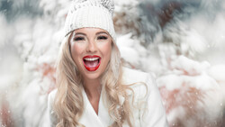 Woman with blonde wavy hair wearing white beanie and coat, smiling with red lipstick in falling snow.