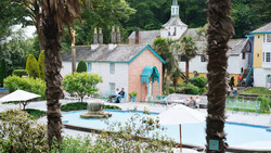 Coastal village with pastel buildings, palm trees framing the composition, and a swimming pool in the foreground.