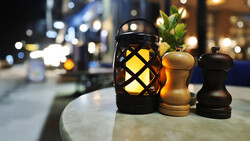 Black lattice lantern with glowing yellow light beside wooden salt and pepper mills on an outdoor table at night.