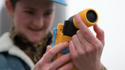 Child holding a yellow and black digital rangefinder device with lens attachment.