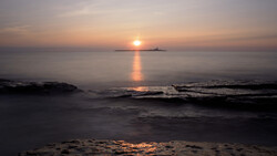 Sun setting over the ocean with a distant island silhouette and golden light reflecting on calm waves.
