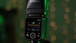V100 SU camera flash with LCD control panel displaying settings against blurred green bokeh background.
