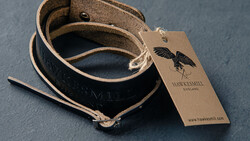 Brown leather camera strap coiled on gray surface with kraft paper hang tag featuring winged logo.