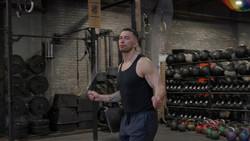 Man in black tank top performing kettlebell exercise in gym with equipment racks visible.