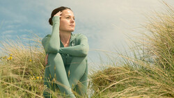 Woman in sage green shirt gazing upward among tall beach grasses under blue sky.