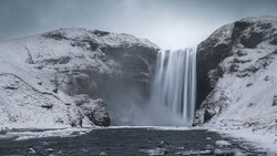 Tall waterfall cascading between snow-covered rocky cliffs into a dark pool of water.