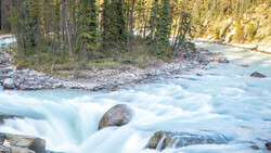 Turquoise river with long-exposure blur flowing around boulders, coniferous forest and rocky hillside in background.