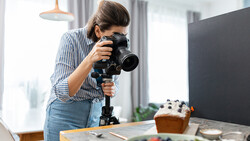 Female videographer operating a camera on a stabilizer rig while filming a styled food scene indoors.
