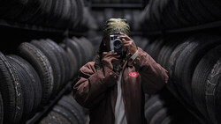 Photographer holding vintage camera surrounded by stacked tires in industrial setting.