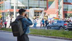 Photographer with backpack shooting an outdoor event with crowds and buildings in background.