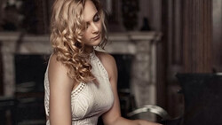 Woman in white lace dress posing in an elegant interior with fireplace backdrop.