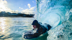 Bodyboarder riding inside a wave barrel with crystalline blue water, photographed from water level with a boat visible in the background.